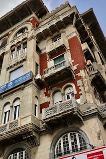 A detailed facade of an old building showcasing intricate architectural elements. The structure features ornate balconies, arched windows, and a combination of red brick and light stone materials. Signs with Arabic script are visible on the building, indicating commercial use. The wear on the facade suggests historical significance.