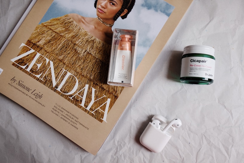 A magazine cover features a person wearing a fringed, brown top with a clear sky in the background. Beside the magazine is a boxed cosmetic product labeled 'Glossier', a green jar labeled 'Cicapair Re-Cover', and a white pair of wireless earphones in their charging case. The objects are laid out on a wrinkled, white surface.