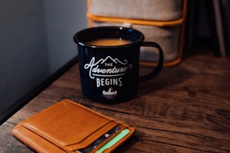 A black mug with the phrase 'The Adventure Begins' written on it sits on a wooden table. Next to the mug, there is a tan leather wallet with several cards partially visible inside.