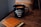 A black mug with the phrase 'The Adventure Begins' written on it sits on a wooden table. Next to the mug, there is a tan leather wallet with several cards partially visible inside.