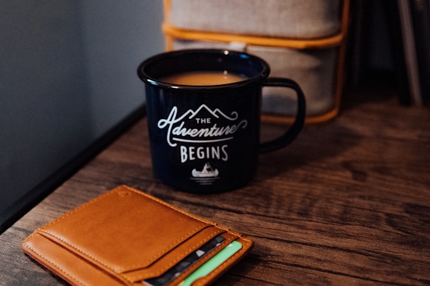 A black mug with the phrase 'The Adventure Begins' written on it sits on a wooden table. Next to the mug, there is a tan leather wallet with several cards partially visible inside.