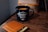 A black mug with the phrase 'The Adventure Begins' written on it sits on a wooden table. Next to the mug, there is a tan leather wallet with several cards partially visible inside.