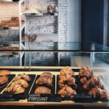 A bakery display featuring an assortment of croissants and pastries arranged neatly on trays. Behind the glass counter, loaves of bread are stacked on shelves against a white brick wall. Labels with prices are visible, offering items such as chocolate croissant, almond croissant, baguette, and white loaf.