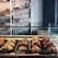 A bakery display featuring an assortment of croissants and pastries arranged neatly on trays. Behind the glass counter, loaves of bread are stacked on shelves against a white brick wall. Labels with prices are visible, offering items such as chocolate croissant, almond croissant, baguette, and white loaf.