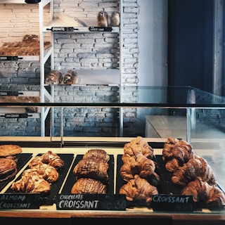 A bakery display featuring an assortment of croissants and pastries arranged neatly on trays. Behind the glass counter, loaves of bread are stacked on shelves against a white brick wall. Labels with prices are visible, offering items such as chocolate croissant, almond croissant, baguette, and white loaf.