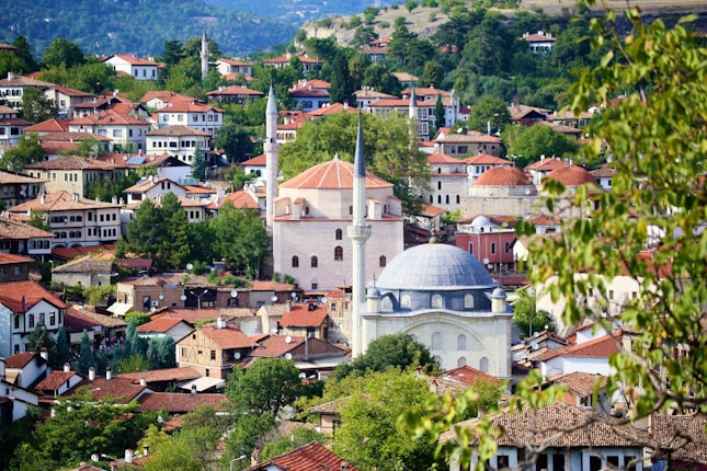 A scenic view of a town with traditional Ottoman architecture, featuring red-roofed houses and green trees. The prominent structures include two mosques with minarets, nestled among the other buildings. The background shows a lush landscape of rolling hills and forests.