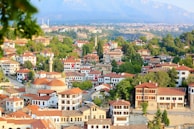 A picturesque town with numerous traditional houses featuring red-tiled roofs, surrounded by lush green trees. Mosques with domes and minarets are visible, adding to the cultural landscape. Rolling hills and mountains form the backdrop, with clear skies overhead. Turkish flags adorn some buildings, highlighting national pride.