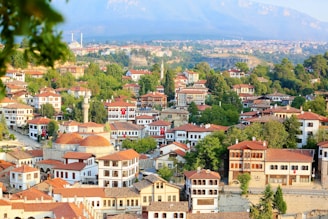 A picturesque town with numerous traditional houses featuring red-tiled roofs, surrounded by lush green trees. Mosques with domes and minarets are visible, adding to the cultural landscape. Rolling hills and mountains form the backdrop, with clear skies overhead. Turkish flags adorn some buildings, highlighting national pride.