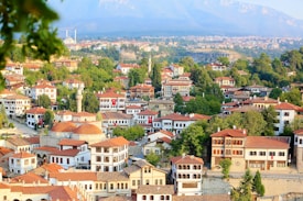 A picturesque town with numerous traditional houses featuring red-tiled roofs, surrounded by lush green trees. Mosques with domes and minarets are visible, adding to the cultural landscape. Rolling hills and mountains form the backdrop, with clear skies overhead. Turkish flags adorn some buildings, highlighting national pride.