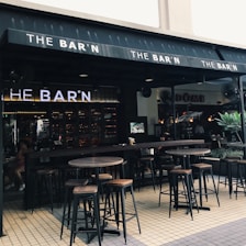 An outdoor seating area of a bar featuring high wooden tables and stools. The bar's name, 'THE BAR'N', is prominently displayed on the awning. The ambiance is relaxed with soft lighting inside. There are fans visible near the entrance, and a few plants adding greenery to the setting.
