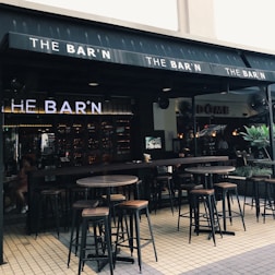 An outdoor seating area of a bar featuring high wooden tables and stools. The bar's name, 'THE BAR'N', is prominently displayed on the awning. The ambiance is relaxed with soft lighting inside. There are fans visible near the entrance, and a few plants adding greenery to the setting.