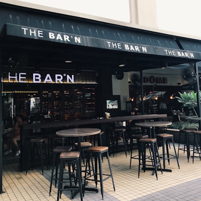 An outdoor seating area of a bar featuring high wooden tables and stools. The bar's name, 'THE BAR'N', is prominently displayed on the awning. The ambiance is relaxed with soft lighting inside. There are fans visible near the entrance, and a few plants adding greenery to the setting.