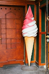 A large, decorative ice cream cone sculpture sits beside a wooden door. The cone features swirled pink and white ice cream on a beige waffle cone base. It is set against a dark green and brown corner.