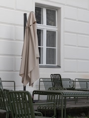 A neatly arranged outdoor seating area features several green metal chairs on a wooden deck, positioned next to a building with a white, paneled wall. An umbrella is folded and placed next to a large window with multiple panes, providing an inviting yet serene atmosphere.