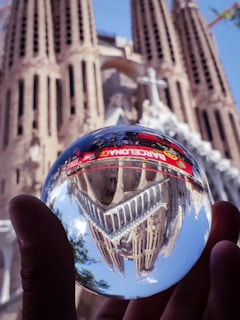 A hand holds a reflective sphere that shows the reflection of a gothic architectural structure with sharp spires. The word 'BARCELONA' is visible on the sphere. The building itself is blurred in the background, with intricate details and a prominent cross.