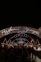 A festive, well-lit Christmas market scene at night with an arched sign displaying '2020' and Cyrillic script. Numerous strings of lights and decorations, including a central Christmas tree, create a warm and inviting atmosphere. People are strolling, wrapped in winter clothing, enjoying the holiday ambiance.