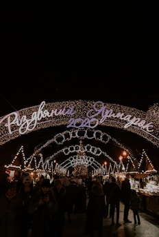 A festive, well-lit Christmas market scene at night with an arched sign displaying '2020' and Cyrillic script. Numerous strings of lights and decorations, including a central Christmas tree, create a warm and inviting atmosphere. People are strolling, wrapped in winter clothing, enjoying the holiday ambiance.