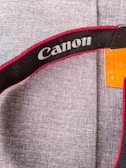 A close-up view of a fabric surface with a camera strap displaying the 'Canon' logo. The strap features a black base with red trim and white text. The background consists of a textured, grey material, and an orange leather piece is partially visible.
