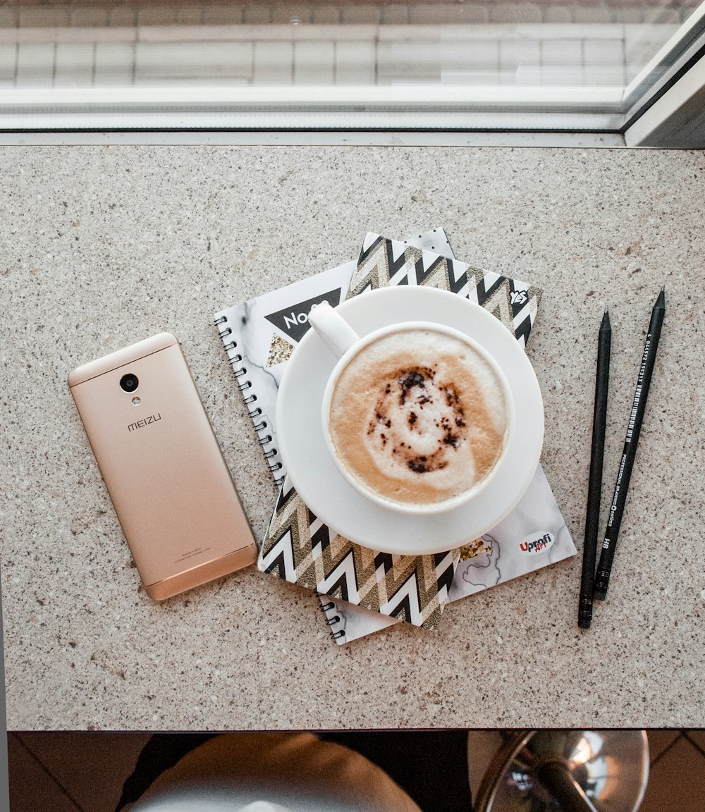 white coffee cup with saucer on notebook between gold Meizu smartphone and two black pencils