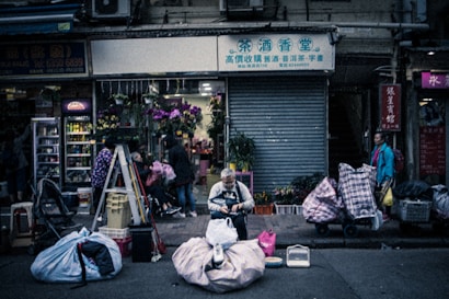 An urban street scene featuring a closed shop with a metal shutter. In the foreground, a person is sitting amidst large bags and various items, appearing to sort through belongings. The scene is busy, with people walking by, and a market-like setup with colorful flowers visible in front of the shop. The environment suggests a mix of commerce and daily activity.