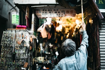 An older man stands in front of a crowded market stall, reaching upward with a long pole. The stall is filled with various items such as belts, watches, and other accessories displayed on racks. Bright lights illuminate the interior, creating a warm glow amidst the busy atmosphere.