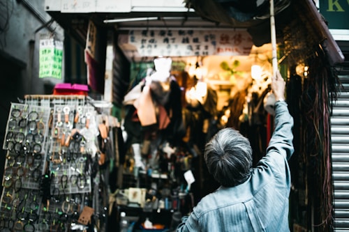 An older man stands in front of a crowded market stall, reaching upward with a long pole. The stall is filled with various items such as belts, watches, and other accessories displayed on racks. Bright lights illuminate the interior, creating a warm glow amidst the busy atmosphere.