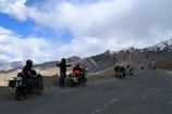 A group of motorcyclists is parked on a mountainous road with snow-dusted peaks in the background. The sky is partly cloudy, and the landscape is rugged and expansive. One of the motorcyclists appears to be gesturing or talking to others.