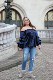A woman wearing a blue off-shoulder top with ruffled sleeves and ripped blue jeans stands on a cobblestone walkway. The background features an ornate building with arched windows and decorative railings. Her shoes are beige ankle boots.
