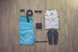 A flat lay arrangement featuring a pair of light blue shorts, a pale pink polo shirt, dark flip-flops with colorful accents, a black belt, a pair of sunglasses, a dark wallet, a wristwatch with a black strap, and a small bottle with a spray nozzle, all placed on a wooden floor.