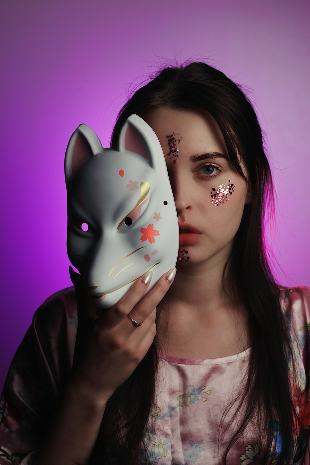 woman holding white and pink cat face mask standing in front of purple background
