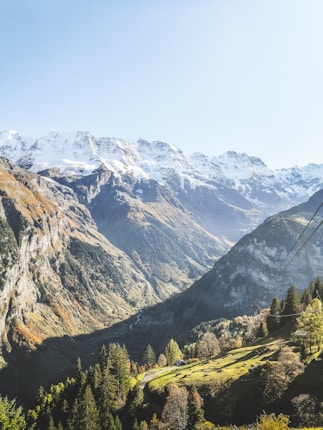 A breathtaking view of a mountainous landscape featuring a series of towering snow-capped peaks. In the foreground, lush green valleys and forested hills provide contrast to the ruggedness of the mountains. The bright sunlight casts distinct shadows, highlighting the textures and depths of the terrain. Sparse clouds appear in the clear blue sky, adding to the serene atmosphere.