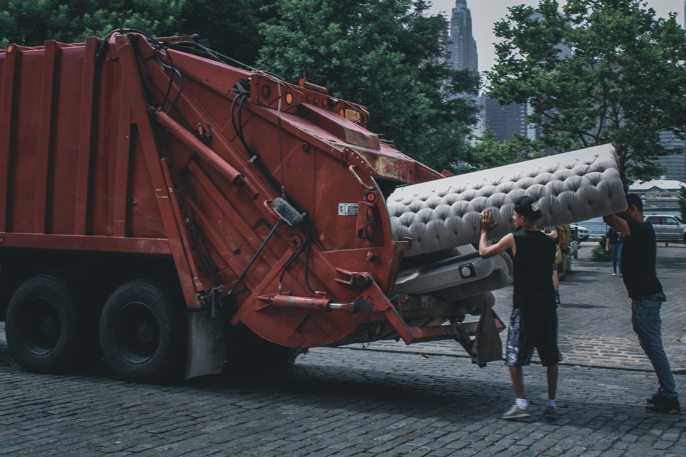 Two workers loading a mattress into a red junk removal truck with a city skyline behind them