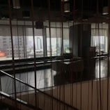 The image captures an indoor space with modern decor, featuring a large window that allows for a view of the distant cityscape. The space includes seating arrangements and some indoor plants, while vertical lines from the design elements add a sense of structure and division.