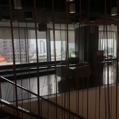The image captures an indoor space with modern decor, featuring a large window that allows for a view of the distant cityscape. The space includes seating arrangements and some indoor plants, while vertical lines from the design elements add a sense of structure and division.