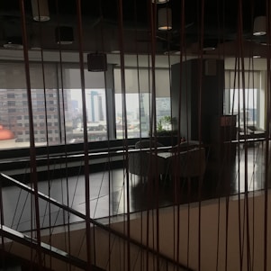 The image captures an indoor space with modern decor, featuring a large window that allows for a view of the distant cityscape. The space includes seating arrangements and some indoor plants, while vertical lines from the design elements add a sense of structure and division.