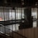 The image captures an indoor space with modern decor, featuring a large window that allows for a view of the distant cityscape. The space includes seating arrangements and some indoor plants, while vertical lines from the design elements add a sense of structure and division.