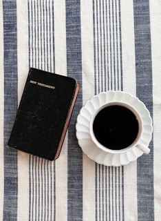 A black New Testament book rests on a striped, blue and white tablecloth beside a cup of black coffee in a white saucer. The scene is simple and neatly arranged, suggesting a moment of calm reflection or a morning routine.