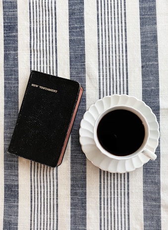 A black New Testament book rests on a striped, blue and white tablecloth beside a cup of black coffee in a white saucer. The scene is simple and neatly arranged, suggesting a moment of calm reflection or a morning routine.