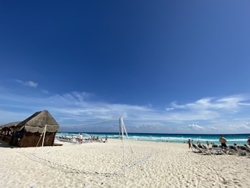 A sandy beach under a clear blue sky with a volleyball net in the foreground. People are lounging on beach chairs and some are walking or playing near the ocean. A thatched hut sits on the left side, providing shade. The ocean is a vibrant turquoise, and the scene conveys a relaxed, leisurely atmosphere.