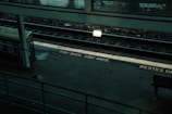 A dimly lit train station platform with a safety message on the ground reading 'STAY BACK. STAY SAFE.' The platform is empty, with tracks visible alongside a bench and a shelter. A sign is illuminated, casting a glow on the tracks and part of the platform.
