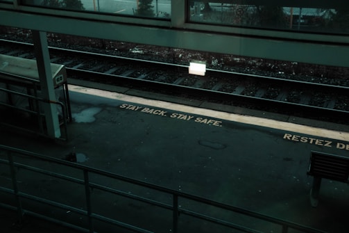 A dimly lit train station platform with a safety message on the ground reading 'STAY BACK. STAY SAFE.' The platform is empty, with tracks visible alongside a bench and a shelter. A sign is illuminated, casting a glow on the tracks and part of the platform.