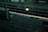 A dimly lit train station platform with a safety message on the ground reading 'STAY BACK. STAY SAFE.' The platform is empty, with tracks visible alongside a bench and a shelter. A sign is illuminated, casting a glow on the tracks and part of the platform.