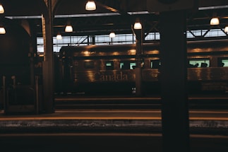 A train station interior with a train on the platform. The station is lit with overhead lamps that cast a warm glow. The word 'Canada' is visible on the side of the metallic train car, and the environment appears dimly lit, suggesting either dusk or an indoor space.