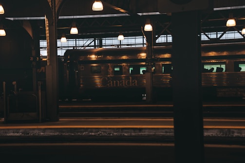 A train station interior with a train on the platform. The station is lit with overhead lamps that cast a warm glow. The word 'Canada' is visible on the side of the metallic train car, and the environment appears dimly lit, suggesting either dusk or an indoor space.