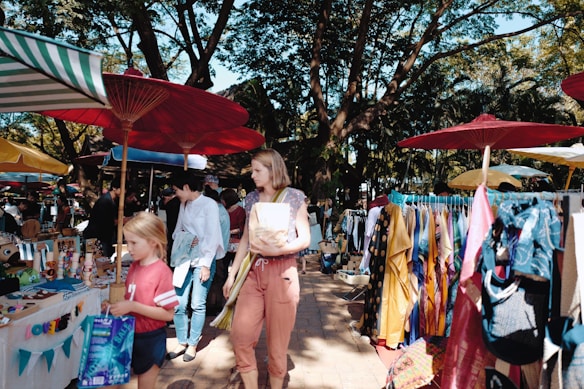 A vibrant outdoor market scene with people browsing colorful stalls under large parasols. Clothing and various goods are displayed alongside the path, surrounded by lush trees providing a canopy overhead.