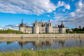 A grand and ornate chateau stands prominently with its elaborate spires and towers, set against a partly cloudy blue sky. A serene moat or river reflects the chateau's image, while lush greenery lines the foreground.