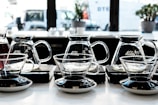 Several glass coffee brewers and cups are aligned neatly on a countertop in a well-lit kitchen or cafe setting. The containers are made of transparent glass, showcasing their elegant design with black and silver bases. The background includes windows that reveal an urban street scene with vehicles and some blurred plants on the windowsill.