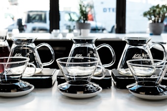 Several glass coffee brewers and cups are aligned neatly on a countertop in a well-lit kitchen or cafe setting. The containers are made of transparent glass, showcasing their elegant design with black and silver bases. The background includes windows that reveal an urban street scene with vehicles and some blurred plants on the windowsill.
