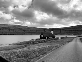 A black and white landscape featuring a small church with a white steeple situated near a body of water. The church is surrounded by a low stone wall and rests on grassy terrain. A winding road runs parallel to the horizon line, leading towards distant cliffs under a cloudy sky.