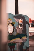 A colorful cartoon standee with cut-out face holes for photography features a person in a hat labeled 'Newport Landing' holding a large fish with a shark in the background. The scene seems humorous and playful, set against a backdrop of blurred outdoor elements.